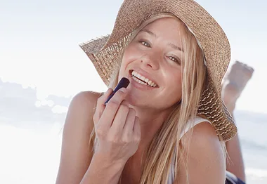 Woman in a sun hat applying lip balm, smiling at the beach.
