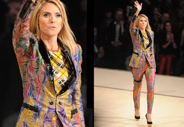 Fashion show runway model in a colorful, patterned suit with a black sequined lapel and high heels, waving to the audience.