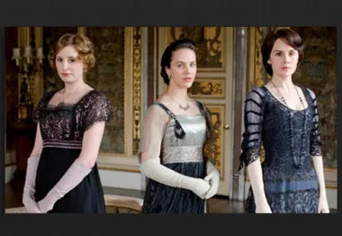 Three women in elegant Edwardian dresses stand in a lavishly decorated room, resembling a scene from Downton Abbey.