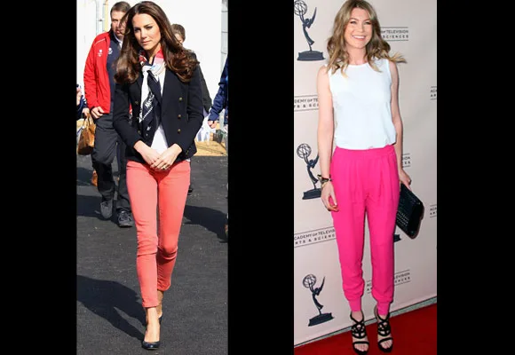 Two women stand side by side wearing coral and pink pants; one in a blazer, the other in a white top, on a red carpet.