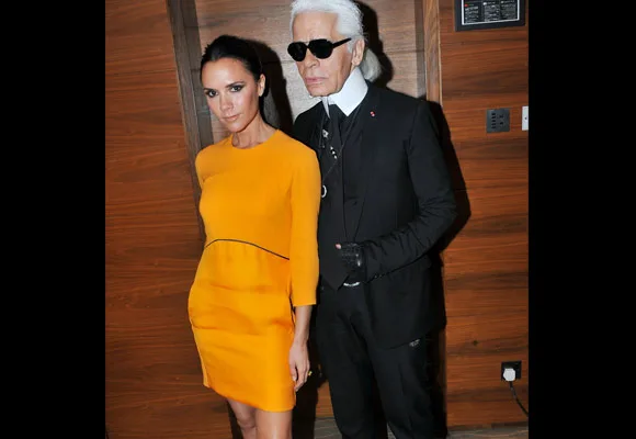 Two individuals standing, one in a bright yellow dress, the other in a black suit and sunglasses, against a wooden wall.