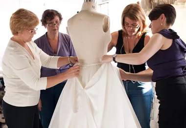 Four designers adjusting a mannequin with a white wedding dress in a studio setting.