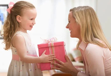 A smiling child gives a pink gift box to a woman inside a home setting.