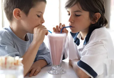 Two children sharing a pink milkshake with straws at a table.