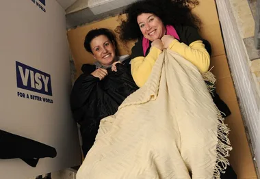 Two women in sleeping bags on cardboard, smiling during the Vinnies CEO Sleepout event next to a Visy box.