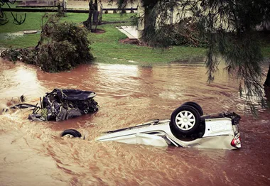 Overturned car submerged in floodwaters with debris, trees in background; scene of Queensland floods.