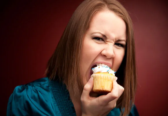 Woman biting into a cupcake with a determined expression against a dark background.