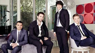 Four men in suits sitting and standing in a modern, stylish room with large windows and artistic decor.