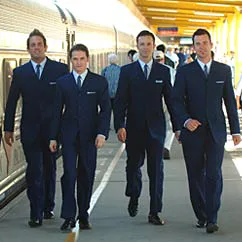 Four men in blue suits walking confidently along a train platform with passengers and a train in the background.