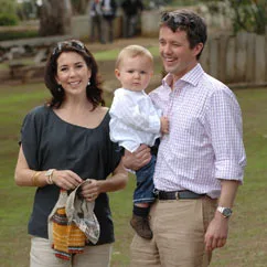 Family smiling and posing outdoors, holding a toddler dressed in a white shirt, casual setting with trees in the background.