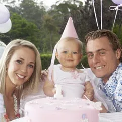 A couple smiles with a baby in a party hat at a decorated outdoor table with a pink cake.