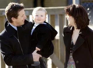 A man and woman smiling at a baby dressed in a dark coat, outdoors near a wooden fence.