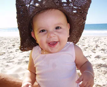A smiling baby wearing a sun hat sits on a beach with the ocean in the background.