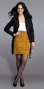 Woman wearing a black coat, white top, mustard skirt, and black heels, holding a clutch against a gray background.