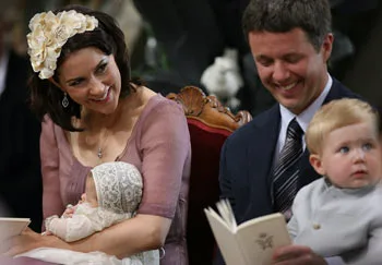 "Family at ceremony, woman holding baby in lace bonnet, man with toddler on lap, both smiling and seated in ornate chairs."