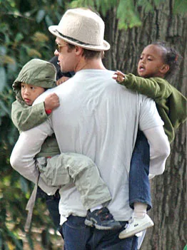 A father wearing a hat carries two children, both in green jackets, outdoors near trees.