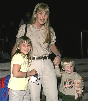 A woman in a khaki outfit stands smiling, holding hands with a young girl and boy; the boy is holding a stuffed toy.
