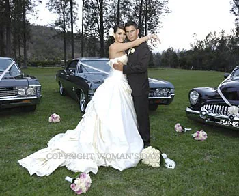 Bride and groom embracing in front of vintage cars on their wedding day, surrounded by trees and flowers.