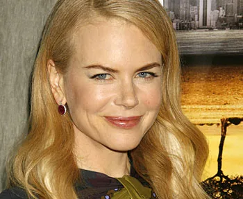 A person with long blonde hair, smiling, in front of a blurred background with yellow and gray tones.
