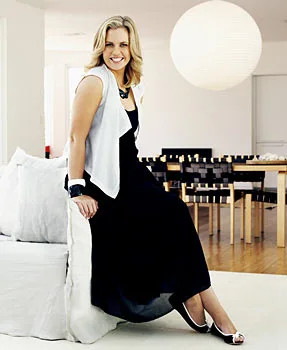 A woman in a black dress and white vest sits on a modern white couch in a stylish, well-lit living room.