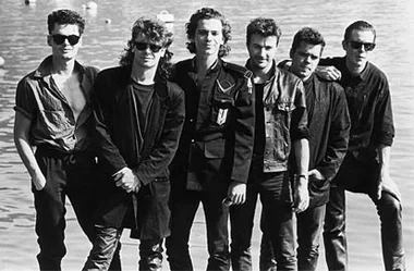 Band members of INXS stand together by a waterfront, dressed in black and wearing sunglasses.