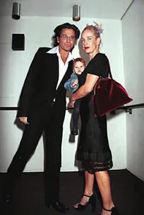A couple poses in formal attire with a toddler in their arms. The woman holds a red handbag.