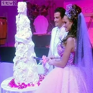 A couple in formal attire cuts a tall, ornate wedding cake decorated with white roses, under pink lighting.