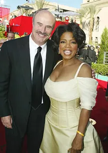 Two people smiling on a red carpet event, one in a black suit and the other in an off-shoulder beige dress.