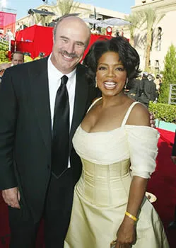 Two people smiling on a red carpet event, one in a black suit and the other in an off-shoulder beige dress.