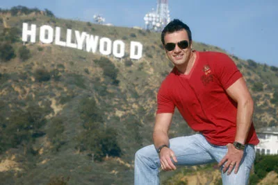 Man in red shirt posing in front of the Hollywood sign on a sunny day.