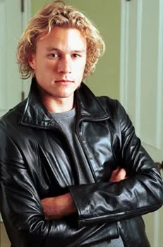Young man with curly hair wearing a black leather jacket, arms crossed, standing confidently indoors.
