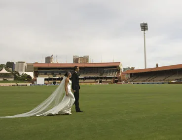 A bride and groom walking on a grassy cricket field with a stadium in the background.