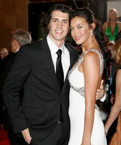 A couple posing elegantly on the red carpet, with the man in a suit and the woman in a white dress.