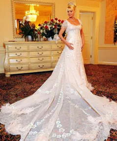 Ivana Trump in an elegant lace wedding gown with a long train, standing in a lavishly decorated room with a dresser and flowers.