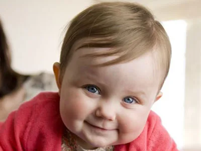 Smiling baby with blue eyes and a pink sweater, leaning slightly forward.