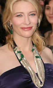 Cate Blanchett at an event, wearing a navy dress and a statement necklace, smiling with a crowd in the background.