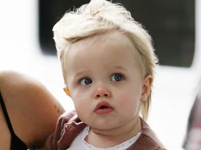 Toddler with blonde hair and blue eyes, wearing a brown sweater, looking curious.