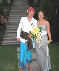 A couple dressed in traditional attire at a wedding, with a staircase and greenery in the background.