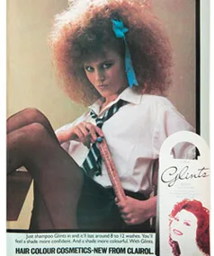 Young woman with curly hair and blue ribbon, sitting with tape measure, wearing school uniform in vintage Clairol ad.