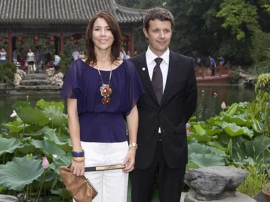 A woman and man stand in front of a pond with green lily pads, wearing formal attire, with a traditional building behind them.