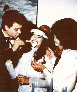 A bride smiles as someone feeds her cake at a wedding celebration, surrounded by people, including a person in a suit.