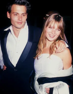 A man in a black suit with a white shirt and a woman in a white dress smiling at an event.