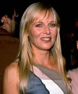 A woman with long blonde hair, smiling, wearing a sleeveless dress in a dimly lit setting.