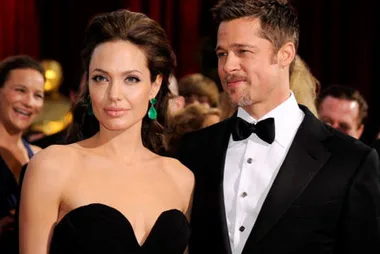 A glamorous couple in formal attire on the red carpet; she wears emerald earrings, he wears a bow tie.