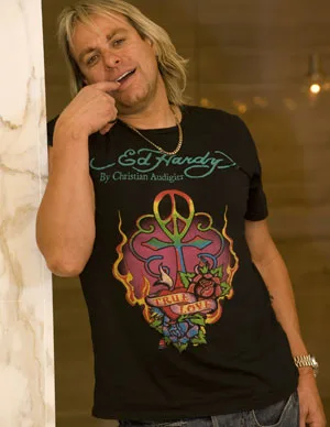 Man with long blonde hair wearing a black Ed Hardy T-shirt, leaning against a marble wall, smiling with a finger on lips.