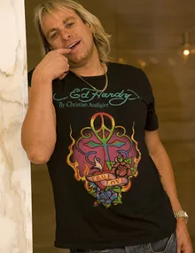 Man with long blonde hair wearing a black Ed Hardy T-shirt, leaning against a marble wall, smiling with a finger on lips.