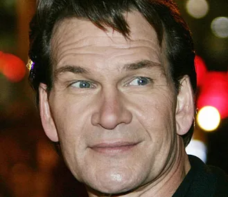 Man with short hair and blue eyes, slightly smiling, in a dark jacket at an evening event with blurred lights in the background.