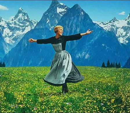 A woman in a dress joyfully twirls in a flower-filled meadow with snow-capped mountains in the background.