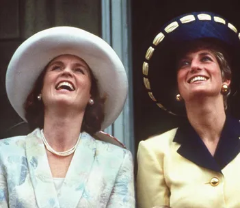 Two women in elegant hats and outfits smiling and looking upwards, one in a white hat and the other in a dark hat with gold trim.
