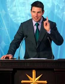 Actor speaking at a podium with Scientology symbol, gesturing with one finger raised.